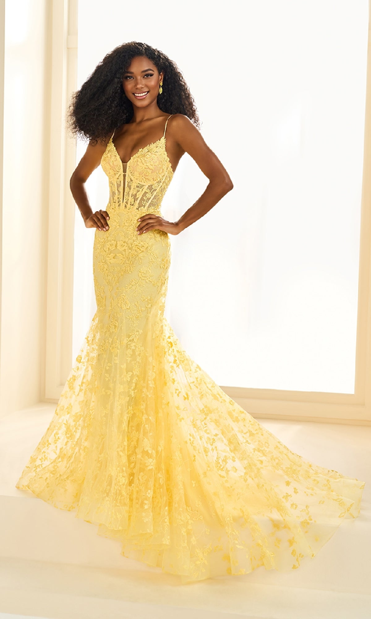 A woman stands in a bright room wearing the Ellie Wilde EW36008, a long embroidered prom dress with thin straps and a sheer, flared train.